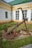 A large, rusty anchor is placed on a concrete base in an outdoor setting. The anchor is surrounded by heavy, rusted chains and is set against a backdrop of a white and yellow building with green trim. There is grass and small patches of vegetation around the base, suggesting a well-maintained garden area.