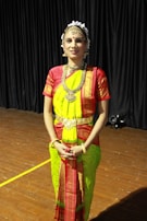 A person is wearing traditional Indian attire, featuring a brightly colored saree in shades of green and red. The outfit includes ornate jewelry, such as a necklace, earrings, and head accessories. The setting appears to be indoors with a wooden floor and dark curtains in the background.