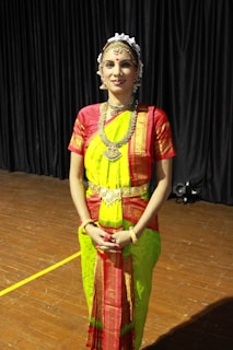 A person is wearing traditional Indian attire, featuring a brightly colored saree in shades of green and red. The outfit includes ornate jewelry, such as a necklace, earrings, and head accessories. The setting appears to be indoors with a wooden floor and dark curtains in the background.