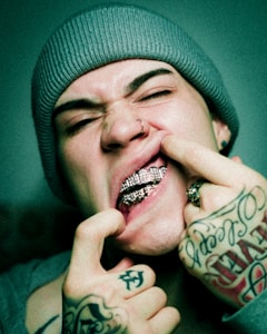 A person wearing a beanie and tattoos on their hands is showing off their diamond-studded grill while pulling their lips back. They have a nose ring and are making a playful, cheeky expression.