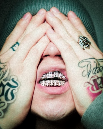 Hands cover a face with tattoos on the fingers. A ring with a decorative design is on one finger. The mouth is open, and diamond-encrusted grills are visible on the teeth, creating a striking visual contrast.