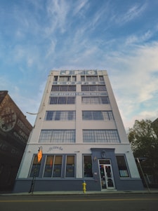 A tall, white building with large windows and signage for JFG Coffee is prominently displayed against a blue sky with scattered clouds. The building features a sign that says 'Jackson Ave' and below it is a bright yellow fire hydrant on the sidewalk.