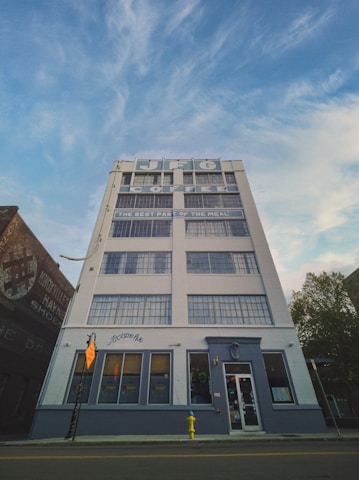 A tall, white building with large windows and signage for JFG Coffee is prominently displayed against a blue sky with scattered clouds. The building features a sign that says 'Jackson Ave' and below it is a bright yellow fire hydrant on the sidewalk.