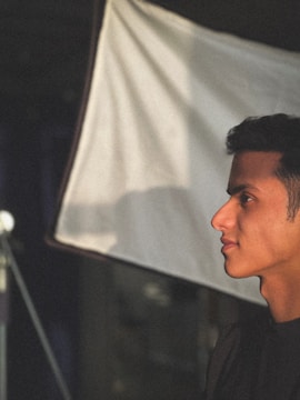 A person is shown in profile view, illuminated by soft lighting, creating a dramatic contrast against a backdrop of a light reflector or screen in a studio setting.