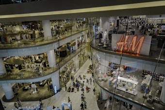 A multi-level shopping mall with numerous stores featuring clothing and accessories. The largest visible store has a prominent H&M sign. People are walking and gathering on the lower floor, giving the area a lively atmosphere.
