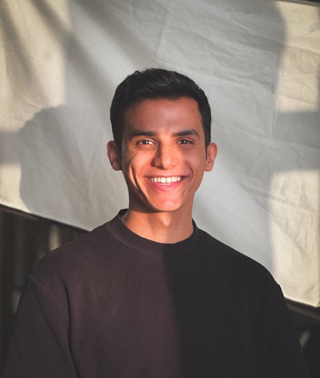A person wearing a dark sweater is smiling warmly while standing in front of a white background with soft shadows cast across it. The lighting creates a gentle contrast on the person's face.