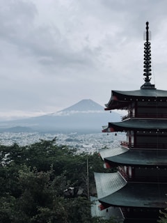 A traditional Japanese pagoda stands prominently in the foreground, with the iconic silhouette of Mount Fuji visible in the background. The scene is enveloped in a moody, overcast sky, adding a sense of tranquility and mystery. Lush green foliage surrounds the structure, and a distant cityscape is faintly visible through the mist.