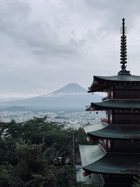 A traditional Japanese pagoda stands prominently in the foreground, with the iconic silhouette of Mount Fuji visible in the background. The scene is enveloped in a moody, overcast sky, adding a sense of tranquility and mystery. Lush green foliage surrounds the structure, and a distant cityscape is faintly visible through the mist.