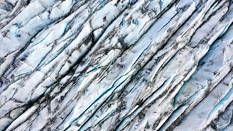 An aerial view of a glacier showing its textured surface with deep crevices and lines formed by years of movement and erosion. The ice appears bluish-white with dark streaks, likely caused by dirt and sediments.