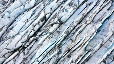 An aerial view of a glacier showing its textured surface with deep crevices and lines formed by years of movement and erosion. The ice appears bluish-white with dark streaks, likely caused by dirt and sediments.