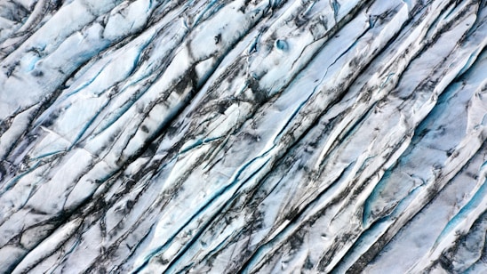 An aerial view of a glacier showing its textured surface with deep crevices and lines formed by years of movement and erosion. The ice appears bluish-white with dark streaks, likely caused by dirt and sediments.