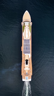 Aerial view of a large cruise ship sailing through the water. The ship has a sleek design with multiple decks, solar panels, and a visible swimming pool at the rear. The vessel is creating wake as it moves forward, and the deep blue water surrounds it.