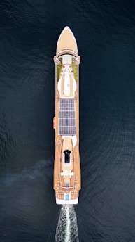 Aerial view of a large cruise ship sailing through the water. The ship has a sleek design with multiple decks, solar panels, and a visible swimming pool at the rear. The vessel is creating wake as it moves forward, and the deep blue water surrounds it.