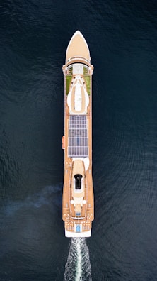 Aerial view of a large cruise ship sailing through the water. The ship has a sleek design with multiple decks, solar panels, and a visible swimming pool at the rear. The vessel is creating wake as it moves forward, and the deep blue water surrounds it.
