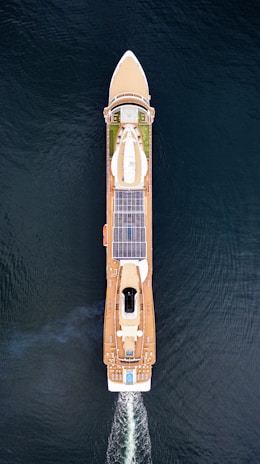 Aerial view of a large cruise ship sailing through the water. The ship has a sleek design with multiple decks, solar panels, and a visible swimming pool at the rear. The vessel is creating wake as it moves forward, and the deep blue water surrounds it.