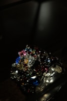 A collection of colorful, multifaceted gemstones is piled together in a dark setting. The gems exhibit a variety of shapes and cuts, reflecting light in different directions. The scene is illuminated to emphasize the brilliance and vibrancy of the stones.