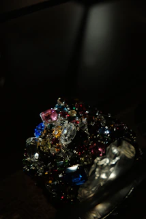 A collection of colorful, multifaceted gemstones is piled together in a dark setting. The gems exhibit a variety of shapes and cuts, reflecting light in different directions. The scene is illuminated to emphasize the brilliance and vibrancy of the stones.