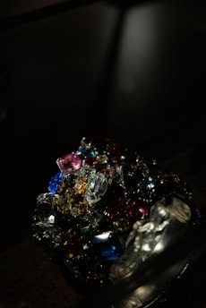 A collection of colorful, multifaceted gemstones is piled together in a dark setting. The gems exhibit a variety of shapes and cuts, reflecting light in different directions. The scene is illuminated to emphasize the brilliance and vibrancy of the stones.