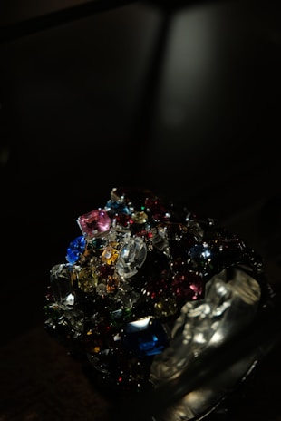 A collection of colorful, multifaceted gemstones is piled together in a dark setting. The gems exhibit a variety of shapes and cuts, reflecting light in different directions. The scene is illuminated to emphasize the brilliance and vibrancy of the stones.