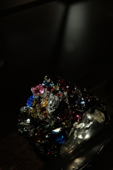 A collection of colorful, multifaceted gemstones is piled together in a dark setting. The gems exhibit a variety of shapes and cuts, reflecting light in different directions. The scene is illuminated to emphasize the brilliance and vibrancy of the stones.