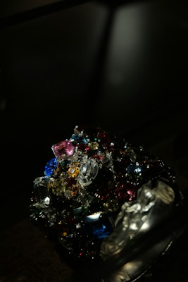 A collection of colorful, multifaceted gemstones is piled together in a dark setting. The gems exhibit a variety of shapes and cuts, reflecting light in different directions. The scene is illuminated to emphasize the brilliance and vibrancy of the stones.