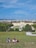 A large, historic palace is situated in the background, with a wide expanse of green garden and a fountain in front. Several people are lounging on a grassy hill in the foreground, enjoying a sunny day under a clear blue sky. The scene is peaceful and picturesque, capturing a moment of relaxation and tourism.