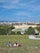 A large, historic palace is situated in the background, with a wide expanse of green garden and a fountain in front. Several people are lounging on a grassy hill in the foreground, enjoying a sunny day under a clear blue sky. The scene is peaceful and picturesque, capturing a moment of relaxation and tourism.