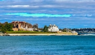 A coastal scene with large, elegant mansions situated on a grassy embankment. The architecture is ornate, with multiple chimneys and gabled roofs, suggesting historic or affluent homes. The nearby ocean is a vibrant blue, reflecting the clear sky partially covered with clouds. Trees and manicured lawns create a serene and picturesque setting.