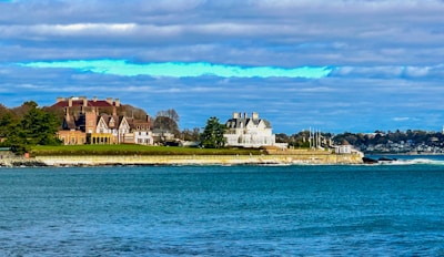 A coastal scene with large, elegant mansions situated on a grassy embankment. The architecture is ornate, with multiple chimneys and gabled roofs, suggesting historic or affluent homes. The nearby ocean is a vibrant blue, reflecting the clear sky partially covered with clouds. Trees and manicured lawns create a serene and picturesque setting.