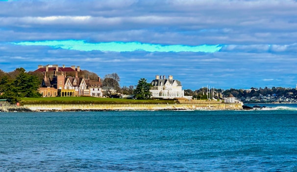 A coastal scene with large, elegant mansions situated on a grassy embankment. The architecture is ornate, with multiple chimneys and gabled roofs, suggesting historic or affluent homes. The nearby ocean is a vibrant blue, reflecting the clear sky partially covered with clouds. Trees and manicured lawns create a serene and picturesque setting.