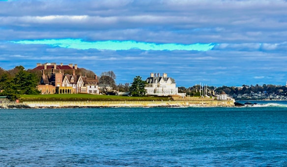 A coastal scene with large, elegant mansions situated on a grassy embankment. The architecture is ornate, with multiple chimneys and gabled roofs, suggesting historic or affluent homes. The nearby ocean is a vibrant blue, reflecting the clear sky partially covered with clouds. Trees and manicured lawns create a serene and picturesque setting.