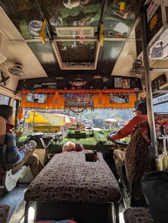 The interior of a bus with two people seated in the driver's and co-driver's seats. The dashboard is decorated with orange garlands and small items. The windshield reveals a view of the outdoors, with tents and parked vehicles visible outside. The bus seats have a patterned fabric, and there are signs of religious or cultural significance displayed.