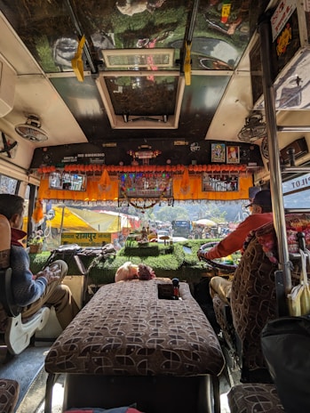 The interior of a bus with two people seated in the driver's and co-driver's seats. The dashboard is decorated with orange garlands and small items. The windshield reveals a view of the outdoors, with tents and parked vehicles visible outside. The bus seats have a patterned fabric, and there are signs of religious or cultural significance displayed.