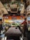 The interior of a bus with two people seated in the driver's and co-driver's seats. The dashboard is decorated with orange garlands and small items. The windshield reveals a view of the outdoors, with tents and parked vehicles visible outside. The bus seats have a patterned fabric, and there are signs of religious or cultural significance displayed.