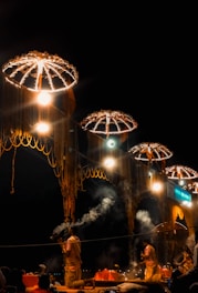 Several people are engaged in a ceremony at nighttime, under large umbrella-like structures decorated with lights and garlands. There are visible plumes of smoke rising from incense. The scene is illuminated by bright lights hanging from the structures.