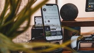 A smartphone stands vertically on a desk, displaying a social media profile. Nearby items include a game controller and a smart speaker, with a blurred plant in the foreground.
