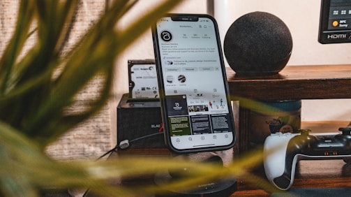 A smartphone stands vertically on a desk, displaying a social media profile. Nearby items include a game controller and a smart speaker, with a blurred plant in the foreground.