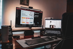 A dimly lit workspace featuring a desktop monitor displaying a website development page and a laptop showing a digital growth insights page. The setup includes a keyboard, mouse, two cups, a pen holder, and a clock displaying '02 36'. The atmosphere suggests a cozy, focused work environment.