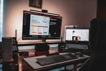 A dimly lit workspace featuring a desktop monitor displaying a website development page and a laptop showing a digital growth insights page. The setup includes a keyboard, mouse, two cups, a pen holder, and a clock displaying '02 36'. The atmosphere suggests a cozy, focused work environment.