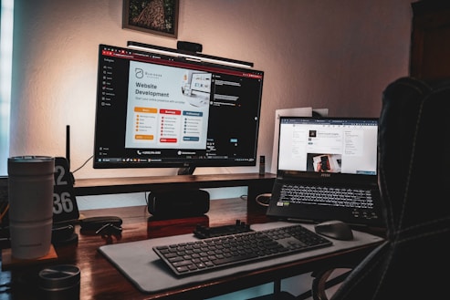 A dimly lit workspace featuring a desktop monitor displaying a website development page and a laptop showing a digital growth insights page. The setup includes a keyboard, mouse, two cups, a pen holder, and a clock displaying '02 36'. The atmosphere suggests a cozy, focused work environment.