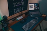 A modern work desk setup featuring a large monitor displaying digital design text, a laptop with an open web browser and music streaming interface, a keyboard, mouse, and a digital clock displaying 5:05. The desk also contains a Bluetooth speaker and an organizer for tech accessories.