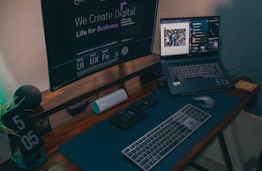 A modern work desk setup featuring a large monitor displaying digital design text, a laptop with an open web browser and music streaming interface, a keyboard, mouse, and a digital clock displaying 5:05. The desk also contains a Bluetooth speaker and an organizer for tech accessories.