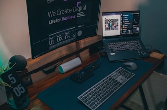 A modern work desk setup featuring a large monitor displaying digital design text, a laptop with an open web browser and music streaming interface, a keyboard, mouse, and a digital clock displaying 5:05. The desk also contains a Bluetooth speaker and an organizer for tech accessories.