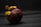 A single mangosteen fruit is positioned against a dark, textured background. The fruit has a round shape with a thick, purple rind and green calyx on top. The lighting creates a dramatic contrast, highlighting the unique texture of the rind and the detailed structure of the calyx.