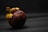 A single mangosteen fruit is positioned against a dark, textured background. The fruit has a round shape with a thick, purple rind and green calyx on top. The lighting creates a dramatic contrast, highlighting the unique texture of the rind and the detailed structure of the calyx.