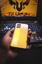 A person is holding a smartphone with a yellow back and a visible camera module. The phone features branding that reads 'realme' and 'DARE TO LEAP'. In the background, there is a laptop with a colorful backlit keyboard and a screen displaying a logo and the text 'TUF GAMING' in bold letters.