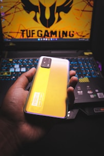 A person is holding a smartphone with a yellow back and a visible camera module. The phone features branding that reads 'realme' and 'DARE TO LEAP'. In the background, there is a laptop with a colorful backlit keyboard and a screen displaying a logo and the text 'TUF GAMING' in bold letters.