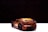 A toy model car is positioned on a flat surface, with a focus on its rear end. The car has a metallic, bronze-like finish with sleek, sporty curves and detailed features like a visible spoiler and black wheels. The background is dark, creating a strong contrast and highlighting the car.