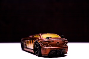 A toy model car is positioned on a flat surface, with a focus on its rear end. The car has a metallic, bronze-like finish with sleek, sporty curves and detailed features like a visible spoiler and black wheels. The background is dark, creating a strong contrast and highlighting the car.