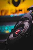 A close-up of a black gaming headset with illuminated red LED accents, placed against a background that appears to be a blurred computer setup with a glowing keyboard.